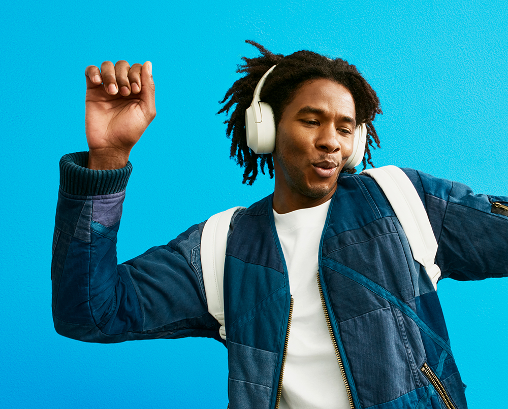 Homem com casaco azul e t-shirt branca, com mochila e auscultadores brancos e os braços elevados, a dançar.